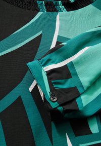 Blusa in tessuto satin con sfondo nero e motivi geometrici turchesi e bianchi. Collo arricciato con dettaglio di polsini bottonati.