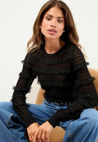 Schwarzer gestrickter Pullover mit Rüschendetails, gestreiftem Muster, rundem Ausschnitt, langen Ärmeln und gerippten Bündchen. Kombiniert mit hochgeschnittenen blauen Jeans.