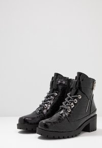 Bottines en cuir verni noir avec motif crocodile, dotées d'œillets métalliques, de lacets noir et blanc, et d'une semelle épaisse avec une fermeture éclair sur le côté.