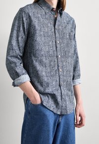 Donkerblauw overhemdbloes met ingewikkelde witte patronen, voorzien van een puntige kraag en opgerolde mouwen. Gepaard met blauwe denim broek.