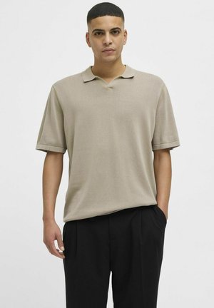 Beige short-sleeve polo shirt with a V-neck and rolled cuffs, featuring a smooth texture and relaxed fit, paired with black trousers.