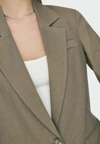 Blazer verde oliva su misura con colletto a vista, chiusura a un bottone e dettagli sulla tasca, indossato su una maglietta bianca. Tessuto morbido.
