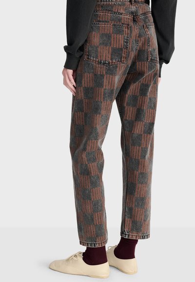 Personne portant un pantalon marron et gris à carreaux avec deux poches arrière, un haut noir à manches longues, des chaussettes bordeaux et des chaussures couleur crème.