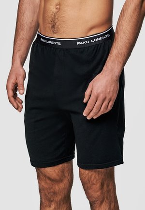 Tors mężczyzny i nogi w czarnych szortach z elastycznym pasem oznaczonym napisem "Pako Lorente" na jednolitym tle.