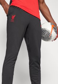 Juodos sportinės kelnės su raudonu „Liverpool FC“ logotipu ir „Nike“ swooshu. Lygus audinys su prigludusia forma, šoninės kišenės ir užtrauktukai ties kulkšnimis.