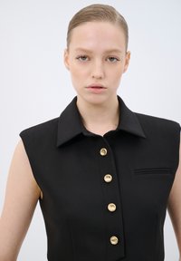 Blusa nera senza maniche con colletto a punta, caratterizzata da cinque bottoni dorati davanti e una piccola tasca sul petto. Tessuto dalla trama liscia.