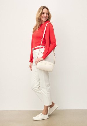 Femeie îmbrăcată într-un tricou roșu cu mâneci lungi și pantaloni albi, purtând o geantă albă de umăr și pantofi sport albi, stând pe fundal simplu.