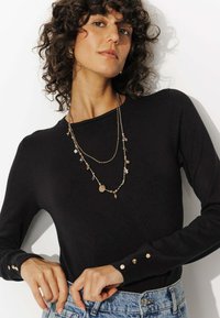 Haut noir à manches longues avec des accents de boutons aux poignets, associé à un collier superposé en ton doré comprenant des perles et un pendentif circulaire.