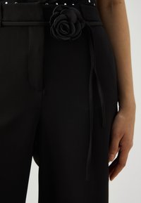 Zbliżenie na czarne spodnie z materiałowym paskiem związanym w kształt kwiatka w talii, a obok luźno oparta ręka.