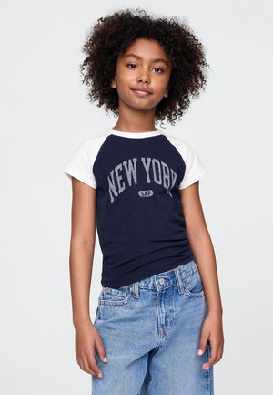Barn med lockigt hår som bär en marinblå och vit "New York"-t-shirt och löst sittande ljusblå jeans mot en enfärgad bakgrund.