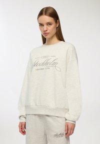 Felpa grigia chiara con scollatura rotonda e polsini a costine, con la scritta "Stockholm Leisure Club" stampata in testo corsivo grigio scuro.