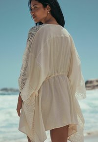 Kaftan bej deschis cu dantelă fină, croială lejeră și talie strânsă, realizat dintr-un material moale și ușor, potrivit pentru plajă.