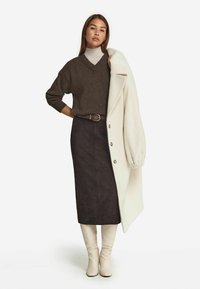 Brauner Strickpullover, beiger Rollkragenpullover, schwarzer Wildledrock und cremefarbener Mantel; beigefarbene kniehohe Stiefel; das Outfit zeichnet sich durch einen Gürtel und Knopfdetails aus.