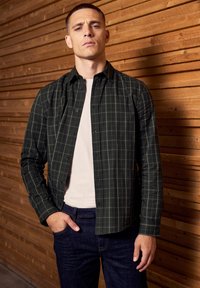 Tmavě zelená károvaná flanelová košile s knoflíky, nošená přes světlejší tričko, doplněná tmavými džíny. Rovný střih a měkká textura.
