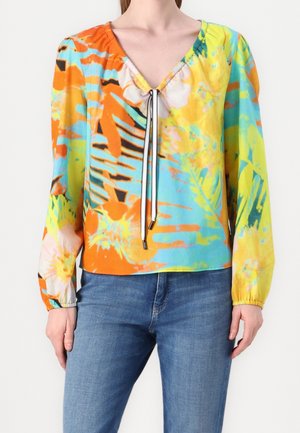 Kobieta nosząca żywą bluzkę z długim rękawem z kwiatowym wzorem tie-dye i sznurkiem z przodu, w połączeniu z niebieskimi dżinsami.