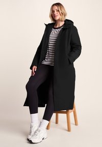 Woman sitting on wooden stool wearing a long black hooded coat, striped shirt, black leggings, white socks, and white sneakers.