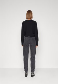 Černý svetr s dlouhými rukávy, zkombinovaný s tmavě šedými džíny. Modelka má na sobě šedé boty na podpatku. Čistý, minimalistický design s uvolněným střihem.