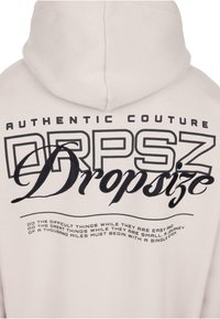 Bluza z kapturem w jasnoróżowym kolorze z czarnym napisem. Zawiera napis "AUTHENTIC COUTURE" oraz stylizowane logo "Dropsize" z motywującymi hasłami poniżej.