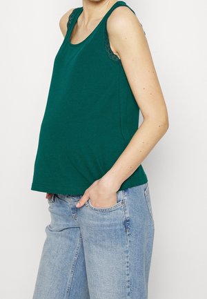 Débardeur vert avec des accents en dentelle sur les épaules, fabriqué en tissu doux. Associé à un jean bleu clair ample, les mains dans les poches.