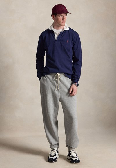 Ung mand iført en burgunderfarvet kasket, navy blå langærmet polo, grå joggingbukser med snøre og sort-hvide sneakers, stående mod en beige baggrund.
