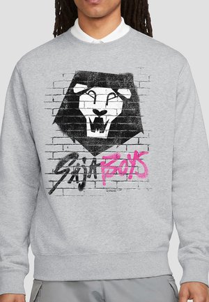 Gray sweatshirt with black and white stylized lion head over brick wall and graffiti-style text "SAGA BOYS" in black and pink.