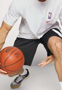 Camiseta blanca de Nike con el logo de la NBA, pantalones cortos deportivos negros y zapatos de baloncesto gris-negros. Sosteniendo un balón de baloncesto naranja.