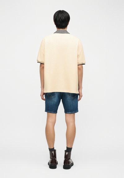 Άνδρας που στέκεται ανάποδα, φορώντας μια μπεζ κοντομάνικη πουκάμισο με καρό γιακά, τζιν σορτς, γκρι κάλτσες και σκούρα καφέ μπότες.