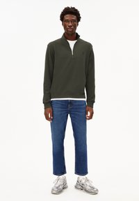 Felpa zip in verde scuro con colletto alto, abbinata a jeans dritti blu e sneakers grigie con dettagli bianchi.