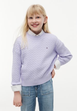 Ragazza bionda sorridente che indossa un maglione lavorato a trecce color lavanda sopra una camicia bianca con le maniche rimboccate e jeans blu, in piedi con la mano sul fianco.