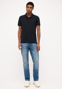 Camisa polo negra con cuello, acompañada de unos vaqueros de mezclilla azul claro. El modelo lleva zapatillas blancas. La tela parece suave y casual.
