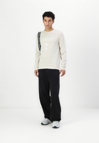 Sudadera de manga larga beige claro con un logo impreso, combinada con pantalones negros y zapatillas grises. La sudadera presenta un detalle de tira negra.