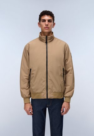 Hombre con chaqueta beige con cremallera, puños y dobladillo elásticos, combinado con vaqueros azul oscuro, de pie sobre un fondo claro y sencillo.