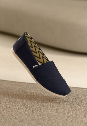 Granatowy but na płaskiej podeszwie typu slip-on wykonany z płótna, z wewnętrzną podszewką w wzór zygzaków (chevron) i beżową podeszwą, pokazany unoszący się nad teksturowaną beżową powierzchnią.