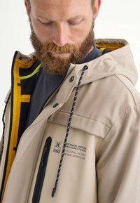 Un homme avec une barbe porte une veste beige avec une doublure intérieure noire et jaune, présentant une poche zippée et des détails de marque.
