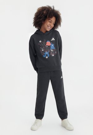 Enfant aux cheveux bouclés souriant, portant un sweat à capuche noir avec des animaux de dessin animé colorés et un pantalon de jogging noir assorti avec des baskets blanches.