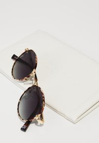 Ochelari de soare cu rame din scoică de țestoasă, cu lentile închise la culoare, accente de metal auriu și un cadru subțire, așezați pe un toc alb texturat marcat cu "le Specs."
