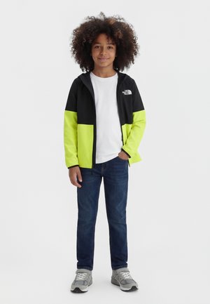 Dziecko z kręconymi włosami ubrane w czarną i neonowożółtą kurtkę, białą koszulkę, niebieskie dżinsy i szare trampki, stojące z jedną ręką w kieszeni.