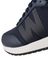Zapatilla azul marino con parte superior de malla y acentos de cuero, diseño de logotipo prominente, suela de goma texturizada y mediopié blanco en contraste.