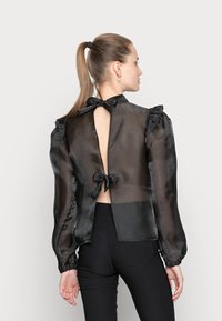 Blusa preta transparente com mangas longas, recorte nas costas, fecho de laço e ruffles almofadados nos ombros. Tecido liso e leve.