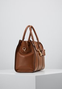Braune Lederhandtasche mit strukturiertem Finish, runder Form, doppelten Griffen, goldenen Hardware-Details und einem kleinen, befestigten Etikett.