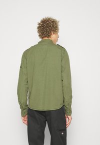 Olive green long-sleeve shirt with a collared neckline and buttoned cuffs. Features shoulder epaulets and a patch on the upper arm.