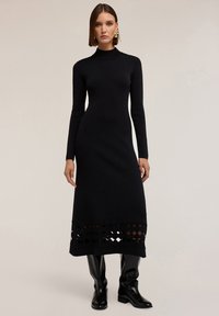 Donna che indossa un vestito midi aderente nero a maniche lunghe con un motivo a diamante tagliato vicino all'orlo e stivali neri al ginocchio.