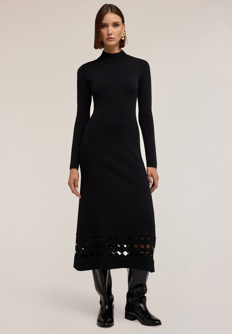 Donna che indossa un vestito midi aderente nero a maniche lunghe con un motivo a diamante tagliato vicino all'orlo e stivali neri al ginocchio.