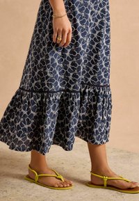 Navyblaues, blumenmusteriertes Maxikleid mit einem rüschengesäumten Saum, kombiniert mit gelben flachen Sandalen mit Zehensteg-Design und verstellbarem Riemen.