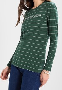 Grünes Langarm-T-Shirt mit weißen horizontalen Streifen, rundem Halsausschnitt und "Calvin Klein Jeans"-Logo auf der Brust.