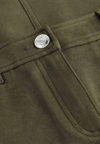 Pantalons en suede verts en gros plan montrant la taille, le bouton en argent, les détails de couture et la passante de ceinture.