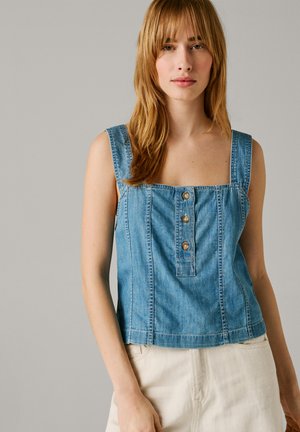 Młoda kobieta o jasnobrązowych włosach, ubrana w bezrękawnik z niebieskiego dżinsu zapinany na guziki oraz jasne spodnie na jednolitym tle.