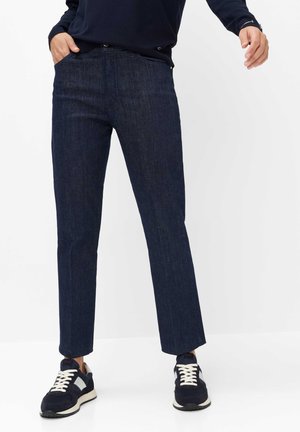 Personne portant un jean droit bleu foncé, des baskets noires et blanches, et un haut à manches longues sombre, debout avec une main dans la poche.