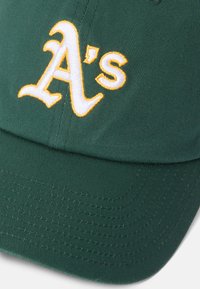 Zelena bejzbol kapa s utisnutim bijelim i zlatnim logom "A's". Tkanina je teksturirana s vidljivim detaljima šivanja na obodu.