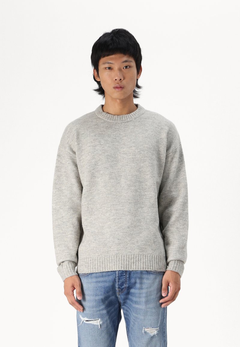 Lichtgrijze gebreide trui met een ronde hals, ribbelmanchetten en een ribbelzoom. Gecombineerd met verweerde blauwe jeans.